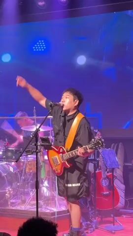 A man in a black shirt and shorts plays a sunburst electric guitar and sings into a microphone on a stage with green and blue lighting, raising his right arm. The camera then shifts to another man, wearing a black t-shirt, shorts, and a backward black baseball cap with a cross necklace, playing a black bass guitar on the same stage under red and blue lighting. A drum set is partially visible in the background.