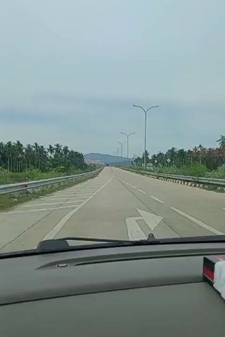The video shows a car driving on a straight, flat, divided highway under a cloudy sky. The highway is lined with palm trees and streetlights. There is a road sign that reads "DILARANG" at the beginning of the clip. The view is from the driver's perspective, focusing through the windshield, with the car's dashboard visible in the foreground.
