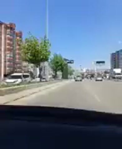 The video shows a view from inside a vehicle, likely a car, moving along a road. The scene is an urban environment with buildings lining the left side of the road. These buildings appear to be residential or office blocks, some with balconies and windows. Trees with green foliage are interspersed between the buildings and along the roadside. The road itself is paved and appears to be a multi-lane highway or a main city road, with other vehicles visible in the distance, indicating traffic. The sky is clear and blue, suggesting a sunny day. The time of day appears to be daytime, likely midday or afternoon, given the bright sunlight and the shadows cast. There are traffic lights and poles along the road. In the distance, there are more buildings and structures, hinting at a larger urban area. The overall impression is of a typical day driving through a city, possibly in Kırıkkale, Turkey, as suggested by the location context. No people are directly visible in the foreground, but the presence of vehicles implies the activity of transportation. There is no discernible text in the video. The mood is neutral, simply documenting a journey through a city.