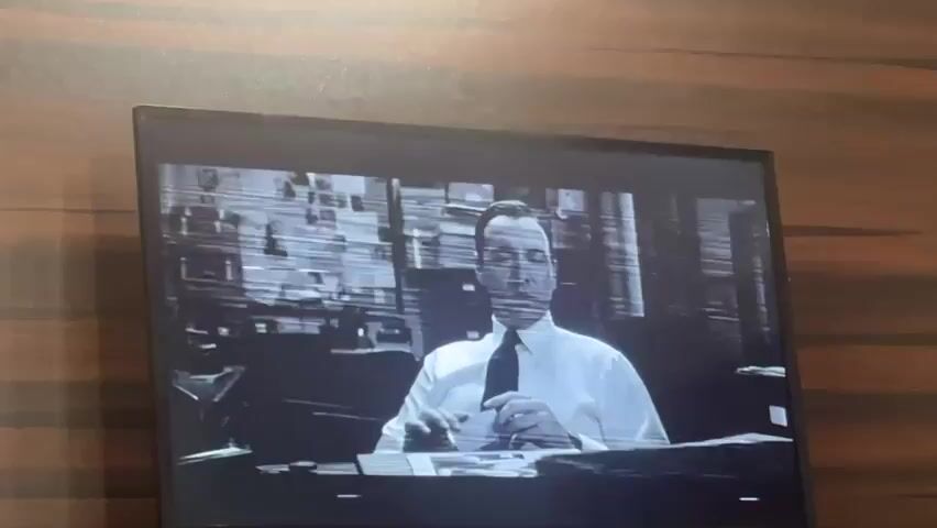 The video shows a static, black-and-white image of a man in a suit sitting at a desk, visible on a television screen. The image has horizontal lines and static interference. He appears to be looking at the viewer. There are objects on the desk, including papers and a small box. The background shows blinds or a window. The TV is positioned against a wooden surface with a light-colored grain pattern.