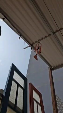 A wind chime, featuring orange cylindrical tubes and a circular top, hangs from a corrugated metal roof or awning. A small, leaf-shaped clapper is visible at the bottom of the chime. Two bare light bulbs are strung along the underside of the same roof structure, with one bulb glowing faintly. The background consists of a clear, bright blue sky on the left and a white building or wall on the right. In the lower left foreground, the top corner of a blue-framed window with small panes is partially visible, while to its right, a section of a brown or orange-framed window also appears. The chimes are gently swaying and producing soft sounds, suggesting a light breeze on a sunny day. No discernible text is visible.