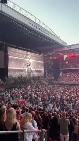 A large crowd of people are gathered in a stadium for a concert. A massive screen displays the performer, who is wearing sunglasses and a black shirt. The stadium is lit with red lights. The crowd is diverse in age and gender, and many people are holding up their phones to record the event. The overall atmosphere appears to be energetic and excited.