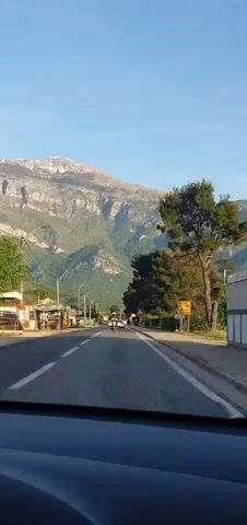 The video provides a first-person perspective from a vehicle driving on a paved road in Kavač, Montenegro, under a clear blue sky. The road, featuring two lanes with a broken white line, extends towards a large, prominent mountain range in the background. The mountains display rocky terrain with patches of green vegetation on their lower slopes and lighter, possibly snow-capped or bare rock, areas near the peaks.

A motorcyclist is visible ahead on the road, with other vehicles in the distance. The immediate surroundings include lush green hills and various trees, notably tall pine-like trees on the right side of the road. Streetlights and power lines run along the left.

On the right side of the road, a yellow rectangular sign is visible, displaying "POTOJ-DOO" and "PUMPA" (which means 'pump'), with "SERVIS" (service) also partially readable below, suggesting a gas station or service area. Further down, a red triangular warning sign with an exclamation mark and a red circular prohibitory sign with a white horizontal bar are visible, indicating road regulations. The bright sunlight suggests it is either morning or late afternoon.