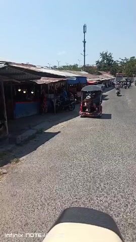The video shows a bright, sunny day with a clear blue sky. The camera is moving along a paved road. On the left side of the road, there are several small market stalls with awnings. People are visible walking and riding motorcycles and a three-wheeled vehicle. On the right side of the road, there is a large building with a red roof, and beyond that, a vacant lot with some construction materials. Power lines are visible throughout the scene, supported by utility poles. The overall impression is of a bustling roadside market and the surrounding environment.