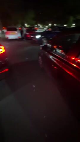At night, a woman sits inside a dark-colored car in a parking lot. The car's brake lights are illuminated, casting a red glow on the interior. The woman looks up, smiles, and then looks down again, seemingly occupied with something on her lap. Other cars are parked nearby under streetlights. The environment appears to be a typical outdoor parking area, possibly near a commercial or recreational site.
