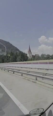 The video is a first-person perspective shot from a vehicle moving along a highway. The view is predominantly of the road ahead and to the side, with a mountain range and trees in the background. A tall, slender red-roofed building, possibly a church steeple, is visible above the trees. A white noise barrier runs along the side of the highway, with metal railings in the foreground. The sky is blue with scattered white clouds. There are reflections visible on the windshield. No people or specific actions are clearly depicted, other than the implied movement of the vehicle.