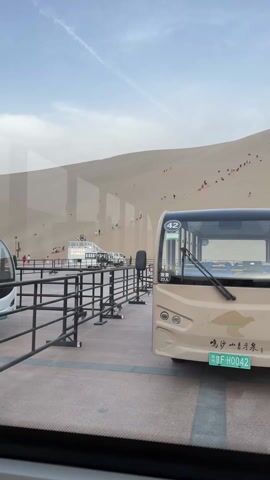 A static, medium shot taken from the inside of a vehicle looks out at a massive sand dune swarming with people. A beige electric bus sits parked in the foreground, facing the dune. The bus is number 42 and has a green license plate that reads "苏HF-H0042". A sign on the bus indicates that it has a speed limit of 30km/h and a capacity of 23 people. Behind the bus, a black metal barrier separates the parking area from the base of the dune. Further back, a second, nearly identical bus is visible. The sky above is pale blue with a few wispy clouds. The dune itself is light beige and its surface is dotted with tiny figures of people walking and climbing. The overall impression is of a tourist destination where visitors are taking advantage of the natural beauty of the landscape.