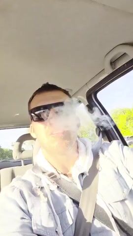 A man is filmed from a first-person perspective inside a car, likely driving. He is wearing dark sunglasses, a light blue denim jacket over a white hooded sweatshirt, and a necklace. His seatbelt is fastened across his chest. The car's headliner, a rearview mirror, and part of the passenger seat headrest are visible in the background.

The man is actively singing or shouting along to music, displaying exaggerated expressions. At first, his mouth is wide open in a yell, then he transitions to a broad, teeth-clenched smile. Wisps of smoke or vapor are briefly visible on the right side of the frame, possibly from a vape or e-cigarette, at the beginning of the clip.

The lighting suggests it is daytime, with bright sunlight visible through the car window. Trees and a blur of scenery can be seen outside, indicating motion. An overlaid text caption reads "DEN STRECkTAN AUF 9 KILOMETER". The overall mood is enthusiastic and energetic.
