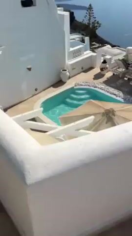 The video shows the exterior of a building with an infinity pool overlooking the ocean. The building is white with classic Greek architecture. The pool is turquoise, with white pebbles decorating the exterior. Two tan umbrellas shade a set of chairs near the pool. Beyond the building is a large body of water with land visible in the distance. A man walks shirtless along the top of the building. The sky is clear and bright blue.