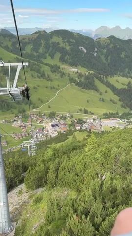 The video begins with a view of a green mountain valley from a moving ski lift. In the foreground, green trees cover the mountainside. Below, a village with red-roofed buildings is nestled in the valley. The ski lift structure, with its metal poles and cables, is visible on the left side of the frame. The camera pans slightly, showing the continued descent of the ski lift cars. The sky is blue with scattered white clouds, and distant mountains are visible in the background. The movement of the ski lift is evident throughout the video.