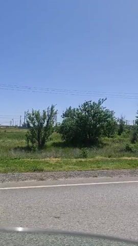 The video, taken from a moving vehicle, shows a roadside scene under a clear blue sky. A damaged utility pole leans dramatically into the adjacent grassy area, supported only by wires at the top. The road surface is asphalt, with green vegetation and trees lining the roadside. A train is visible in the background, moving parallel to the road.  The camera moves steadily forward, maintaining a consistent view of the environment.