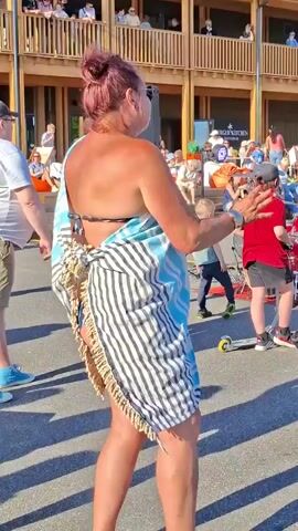 The video shows a woman from behind, wearing a striped sarong tied around her body over a black bikini top and bottoms. Her hair is pulled up in a bun, and she is wearing sunglasses. She is standing outdoors in what appears to be a public space with other people in the background, some of whom are seated. A boy in a red shirt and black shorts is visible to her right, as is a drum set and a scooter. The woman's sarong is open at the back, revealing her lower back and buttocks. The background shows a wooden building with balconies and people on them, suggesting an outdoor gathering or event. The lighting indicates it is daytime.