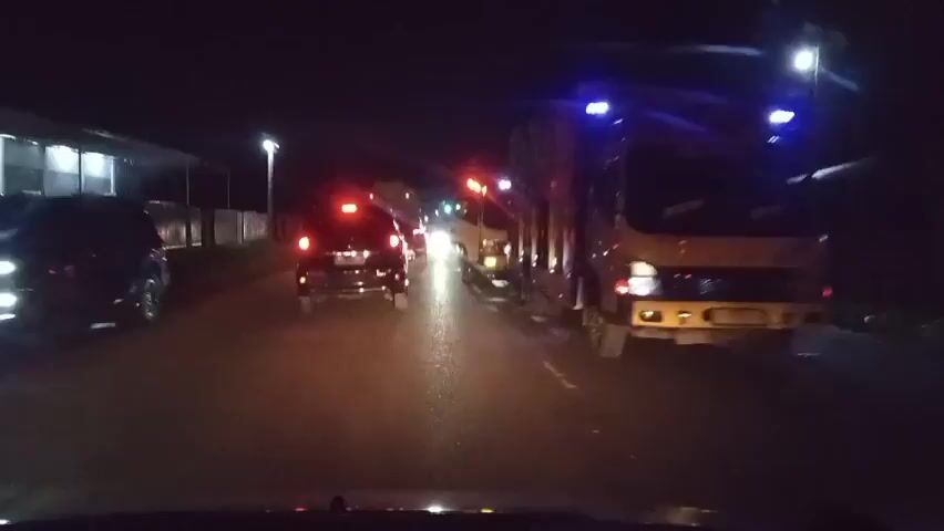 The video shows a nighttime view from inside a vehicle moving along a road. Multiple vehicles are visible, including a black car ahead, a yellow truck to the right, and other vehicles and motorcycles further down the road. The road appears wet, reflecting the lights of the vehicles and streetlights. Some vehicles have their hazard lights or emergency lights flashing, indicated by red and blue lights. The environment is dark with limited visibility, and structures resembling buildings or sheds can be seen along the left side of the road. The overall scene suggests traffic on a road at night, possibly with some form of temporary obstruction or incident causing a slowdown.