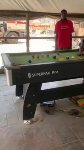 The video shows two men playing pool in an outdoor, covered space. The pool table is black and labeled "SUPERMAX Pro." One man, wearing a red shirt, stands by the table, holding a pool cue. The other man, wearing a dark shirt with a print, takes a shot. Pool balls are scattered across the green felt of the table. The floor is tiled. A bus is visible outside the covered space. Various items, including PVC pipes, a woven rug, and debris, are located around the pool table.