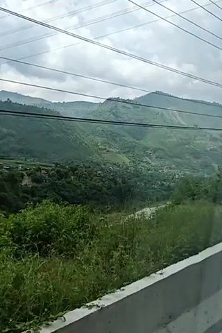 A landscape view of a valley with lush green hills and a river. There are power lines in the foreground and the sky is cloudy. The video pans slightly to the left, revealing more of the valley and the river. There are some houses visible in the distance, nestled in the hills. The vegetation is dense and appears to be natural, with some terraced fields also visible. The overall impression is one of a rural and scenic location.