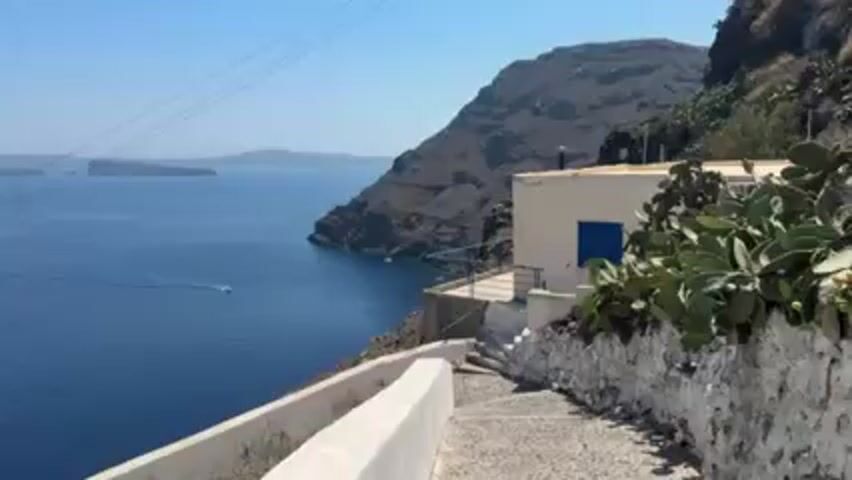 The scene depicts a view from a high vantage point on the Greek island of Santorini. The foreground features a white stone pathway with steps descending along a cliffside. To the right of the pathway is a small, white building with a blue door, surrounded by lush green cactus plants. A metal railing encloses a small terrace in front of the building.  The backdrop showcases the deep blue Aegean Sea, dotted with small boats leaving wakes.  In the distance, a volcanic island rises from the sea, and the sky above is clear and bright blue. Power lines are visible diagonally crossing the top left corner of the frame. The image conveys a tranquil, sunny day in a beautiful Mediterranean setting.