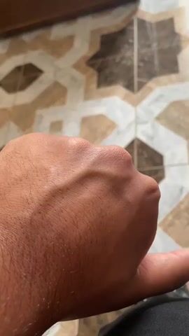 The video shows a close-up view of a person's hand, with prominent veins visible on the back of the hand. The hand is clenched into a fist, with the thumb extended downwards. The background appears to be a tiled floor with a patterned design. The lighting is somewhat dim, casting shadows on the hand and the floor. The focus is on the texture and details of the skin and the raised veins.