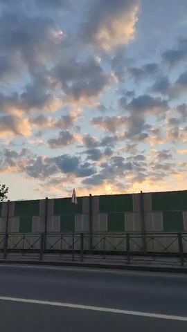 The video pans to the right across a scene dominated by a sky filled with clouds, illuminated by a sunset. In the foreground, a road is visible, with a metal fence separating it from the area beyond. Behind the fence, there are several trees, a large sound barrier, a building with the sign "АВТОСАЛОН КИТ" (Car Dealership KIT), and a parking lot full of cars. A street lamp is also present. The overall impression is of an urban or suburban setting at dusk.
