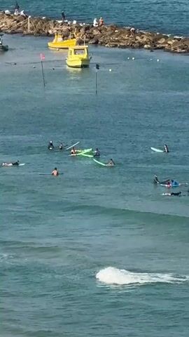 The video shows a beach scene with several people on surfboards in the ocean. There are a few boats anchored in the background near a rocky breakwater. The water is blue and appears to be calm with small waves. Some people are lying on their surfboards, while others are sitting or paddling. In the distance, on the shore, there are more people and what appears to be beach chairs. The overall impression is of a recreational activity at the beach.
