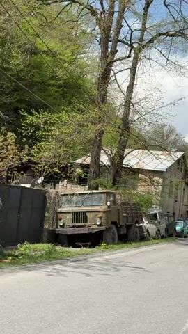 The camera pans slowly across a scene of an impoverished rural area. A large, old, olive-green truck with a flatbed is parked on the shoulder of a paved road. Several old, similarly aged and deteriorating cars are parked in a row behind it, lining the street. Behind the vehicles is a faded, two-story building with multiple windows. The building appears to be in a state of disrepair, with visible signs of aging and weathering on its exterior. The trees are beginning to sprout leaves, indicating it is likely springtime. The backdrop is a hillside covered with lush green vegetation. As the camera continues to pan, a small, makeshift food stall with a green mesh screen comes into view. The stall is followed by more cars, also showing signs of age and disuse. Beyond the stall, further up the road, is a vehicle that appears to be a tank. The sky is partly cloudy, providing a moderate amount of light to the scene.