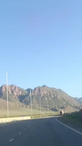 The video shows a view from a vehicle moving on a paved road. The road curves to the right. In the distance, a red semi-trailer truck is visible driving on the same road. Mountains with rocky, brown, and green slopes form the backdrop under a clear blue sky. Several tall poles line the side of the road. The terrain on the left appears to be a flat, grassy valley. The sun is shining, indicating daylight.
