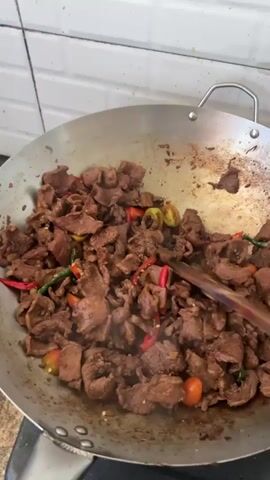 A close-up view shows a large metallic wok on a stovetop, filled with pieces of stir-frying beef, red chili peppers, green chili peppers, and small round red and yellow vegetables, possibly cherry tomatoes or small bell peppers. A wooden spatula or stirrer is actively mixing the ingredients in the wok. The beef pieces are irregular in shape and appear well-browned. The chili peppers are scattered throughout the dish, adding vibrant color. The background features a white tiled wall, typical of a kitchen setting. The activity is the preparation of a meal, likely a stir-fry, in Sidomulyo Barat, Indonesia. No specific text is visible.