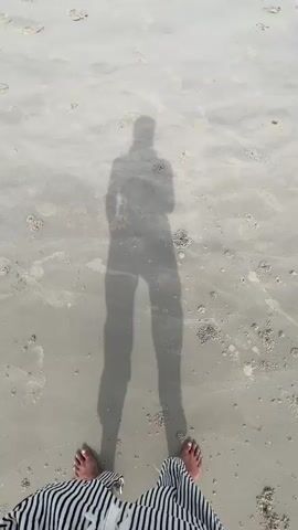 The video shows a person's feet and a shadow of a person standing on a sandy beach. The sand is light-colored with small shells and debris scattered across it. The shadow of the person is long and distinct, with visible legs and torso. The person's feet are visible at the bottom of the frame, wearing sandals or barefoot, with striped fabric on their legs. The camera angle is looking down at the sand and the shadow. There are no other people or significant objects visible in the frame.