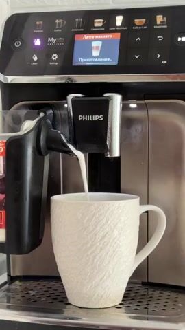 The video shows a Philips coffee machine in action. It is dispensing a creamy white liquid, likely milk, into a textured white mug. The coffee machine's display screen shows "Latte Macchiato" and "Preparing..." with an icon of a layered coffee drink. The coffee machine has a black and silver finish. The mug is positioned on a drip tray. The lighting appears to be natural, coming from above and slightly to the right. The overall environment seems to be a kitchen or an office break room. The video is a close-up shot, focusing on the coffee preparation process.