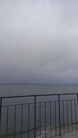 The video shows a choppy sea under a cloudy sky. A dark railing runs across the foreground, obscuring the bottom portion of the frame. A small island or landmass is visible in the distance on the horizon. There are no people or discernable actions taking place.