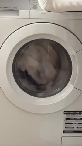 This is a close-up video of a white washing machine. The washing machine door is open, and laundry is inside the drum. The laundry appears to be a mix of white and dark-colored clothing. The drum is not spinning, indicating the washing cycle is not in progress. The top of the washing machine has some text and controls visible, along with a white object placed on top. A sticker with German text, possibly a service offer or warranty information, is adhered to the lower right side of the washing machine door. The lighting is consistent, suggesting an indoor environment.