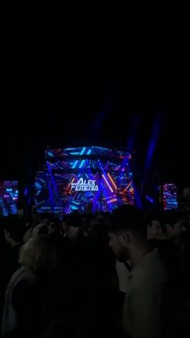 This video shows a crowd of people gathered at an outdoor event at night. A large stage with a brightly lit LED screen is visible in the background, displaying the name "ALEX FENEMIA" in large letters. The screen is illuminated with vibrant colors and geometric patterns. On the left side of the screen, there are smaller LED panels showing similar colorful visuals. Fireworks are set off in the air, creating trails of light and smoke above the crowd. The people in the foreground are mostly silhouetted against the stage lights and fireworks, but some faces and details of clothing are discernible. The atmosphere appears to be festive and energetic.