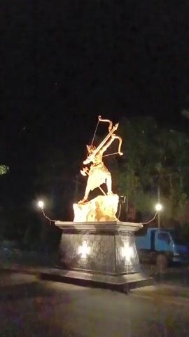 The video shows a golden statue of a warrior with an axe and bow, positioned on a rocky base atop a dark gray plinth. The statue is illuminated against the night sky by lights surrounding the base. In the background, there is a dimly lit street scene with some trees and a blue truck parked nearby. The camera zooms in closer to the statue.