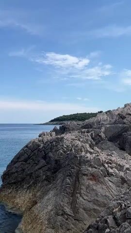 This is a vertical video of a rocky coastline meeting a calm, clear blue sea under a bright blue sky with scattered white clouds. The sun appears to be high in the sky, suggesting it is daytime, likely midday or early afternoon. The weather appears pleasant and clear.

The foreground is dominated by a rugged, grey and brown rocky shore, with waves gently lapping at the base of the rocks. The water is a vibrant turquoise and blue, becoming a deeper blue further out towards the horizon. In the distance, a faint, green, tree-covered headland or island is visible on the right side of the frame, curving around to meet the sea. Further in the distance, on the left side, a hazy line of mountains can be seen on the horizon. There are no people or specific objects clearly identifiable in the video, focusing entirely on the natural landscape. The overall mood is serene and peaceful, showcasing the beauty of a coastal environment. There is no visible text in the video.