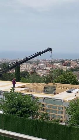 A crane is lifting a large glass panel onto the roof of a building under construction. A person is standing on the roof, directing the crane operator. The building appears to be a modern home with a wooden frame and large windows. The construction site is located on a hillside overlooking a city and the ocean. The sky is overcast.