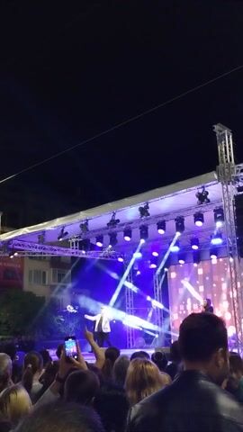 The video captures an outdoor night concert or performance from within the audience. A large, elevated stage dominates the right side of the frame, brightly illuminated with numerous blue and purple stage lights and white spotlights that cast dynamic beams across the scene. Behind the performers, a large LED screen displays vibrant, abstract patterns. At least two individuals are visible on stage; one performer in a white top is centrally located, seemingly addressing or performing for the crowd, while another figure is further back.

The audience, seen from behind and from a slightly elevated perspective within the crowd, occupies the lower right portion of the frame, facing the stage. Several audience members are holding up smartphones, recording the event. The atmosphere is energetic, highlighted by the dynamic lighting and the presence of live music.

In the background, beyond the stage, a building with windows is visible, suggesting an urban setting. Partially visible Cyrillic text on a sign on this building reads "... ИЗРАБКА," which aligns with the provided context of Yambol, Bulgaria. Overhead, power lines or cables stretch across the dark night sky. The video is shot vertically, providing an immersive view of the performance from the audience's perspective. The audio features upbeat music with a driving rhythm and melodic elements, accompanied by sounds from the attending crowd.
