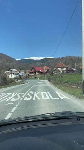 The view from inside a car driving slowly towards a curve in a rural road, on a bright sunny day. In the distance, snow-capped mountains are visible. The road is paved and marked with zebra crossing lines and "ŠKOLA" written on the asphalt, indicating a school zone. Several houses with various architectural styles are scattered along the roadside, some with red roofs and wooden fences. Utility poles and wires run along the road. The windshield is visible, with some dirt or dust present. There are no people visible in the video.