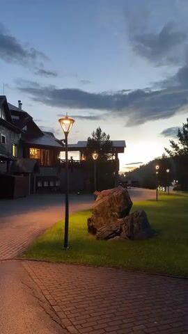The video shows a paved walkway bordering a grassy area with a large rock and several trees. In the background, a building with illuminated windows is visible, as are lampposts and more trees lining a road. The sky is a gradient of blue and gray, with clouds suggesting either dawn or dusk. The lighting is dim, with the lampposts casting yellow light onto the grass and walkway. There are no people or animals visible in the video.