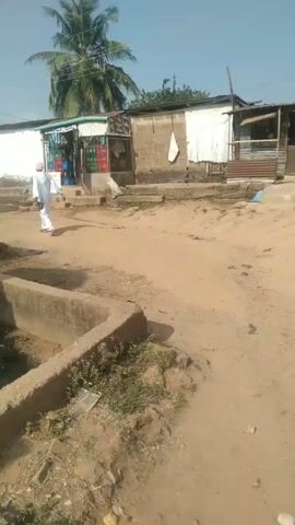 A person in a white robe and pink head covering walks down a dirt road in front of a white car in a rural area. There is a concrete drainage ditch to the left and a small shop selling drinks in the background. The road is rough and dirty, with scattered trash and vegetation. There is a palm tree visible and other buildings in the background. The day is sunny.