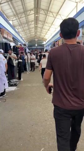 The video shows a person walking through an outdoor market. The market appears to be covered by a metal roof, and stalls line both sides of a central walkway. The stalls display clothing and other goods. Several people are walking in the market, including men, women, and children. The person filming is walking away from the camera, towards the end of the market. The camera then pans to the right, showing a woman with a shopping cart full of bags. The video ends with the person filming continuing to walk through the market.