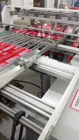 This media file captures a close-up view of an industrial machine in operation, likely within a manufacturing or printing facility. The primary subject is a large, automated machine with various metal components, including rails, rollers, and what appears to be a conveyor system.

The machine is processing sheets of material covered in a repeating red pattern with white text or graphics. These sheets are being fed through the machine, suggesting an automated process like printing, cutting, or packaging. The setup indicates a high-volume production environment.

Notable details include the bright lighting, characteristic of an indoor industrial setting. The specific location cues provided ("Meget, Russia") suggest this is a Russian industrial site. There is no indication of the time of day or weather conditions as the scene is entirely indoors. No people are visible in the frame, emphasizing the automated nature of the activity. The overall tone is one of efficient, mechanical production.

Visible text includes a red label with white text that reads "ОПЕРАТ" which is likely a partial word in Russian, possibly "ОПЕРАТОР" (Operator) or "ОПЕРАЦИЯ" (Operation), further reinforcing the context of a working machine. The sheets themselves have a repeating design, with visible white elements that could be text or graphics, though the details are too small to decipher clearly.