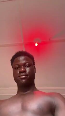 The video shows a shirtless young man looking up at the camera. He has dark skin and short, dark hair. The room he is in has a red light on the ceiling. The walls are orange. There are pictures on the wall behind him. The camera is angled upwards.