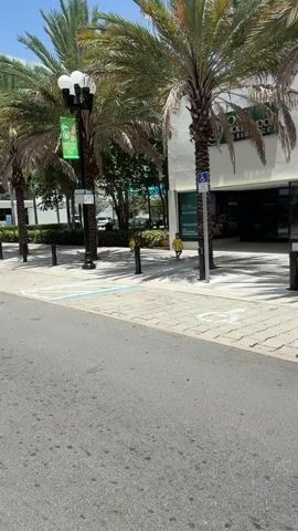 The video pans across a street scene. In the foreground, there is asphalt. To the left of the asphalt, there is a sidewalk with interlocking pavers. Several palm trees are visible. Behind the sidewalk, there is a white building with large glass windows and doors. A sign is visible above the entrance, partially obscured by a palm tree. A blue and white handicapped accessibility symbol is mounted on a pole near the entrance. Further down the sidewalk, a black trash receptacle stands next to a light pole. To the right of the building, a large purple bus with the words "Hard Rock" visible is parked. The bus has an open side, revealing a black interior. Several black speaker-like objects and orange traffic cones are placed around the bus. People are walking on the sidewalk and street, some in yellow shirts. The weather appears to be sunny with some clouds.