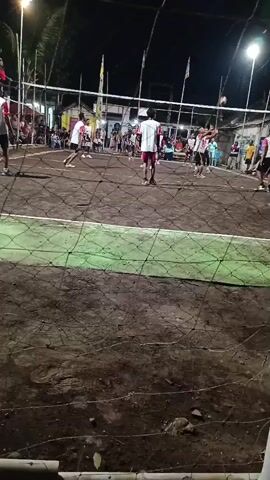 A group of young men are playing volleyball on a dirt court at night. The court is lit by overhead lights. The players are wearing casual clothing. The game appears to be competitive, with players diving and jumping to hit the ball. A crowd of spectators is watching the game from the sidelines. A net divides the court. The court appears to be outdoors, with trees visible in the background. The overall atmosphere is lively and energetic.