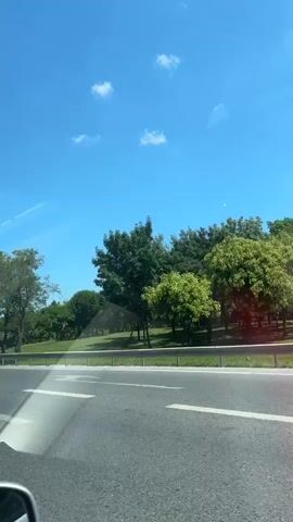 The video captures a roadside view from a moving vehicle on a clear, sunny day. The sky is a vibrant blue with a few scattered white clouds. Trees line the side of the road, some with full green foliage and others with lighter, feathery leaves. A metal guardrail separates the road from a grassy area. In the distance, a Ferris wheel is visible, peeking above the trees, suggesting the presence of an amusement park or similar attraction. A white flatbed truck with a license plate that reads "34 MJB 211" is seen in the foreground. Road signs, including one with Turkish text and an arrow pointing right, are also visible. The road itself has clear lane markings.