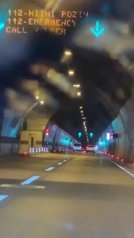 The video shows a car's perspective driving through a tunnel. The tunnel has a high, arched ceiling with rows of lights illuminating the path. The walls of the tunnel are lined with reflective panels, some of which are lit with green and red lights, likely signaling traffic flow or lane status. Several other vehicles, including a white van and cars, are visible ahead, also traveling through the tunnel. There is some mist or fog present, creating a slightly hazy effect and obscuring the view in the distance. The road surface is paved and marked with white lines. The overall impression is of a modern, well-lit tunnel with active traffic.