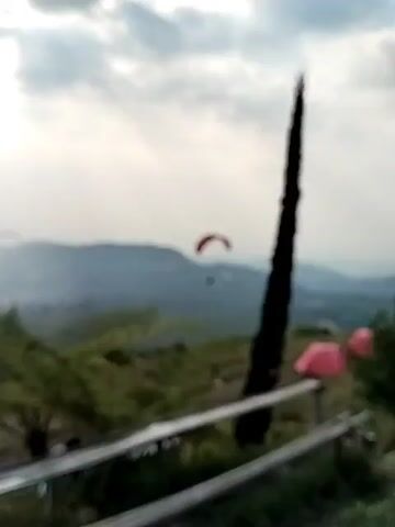 A red paraglider is seen flying in the sky above a mountainous landscape. The paraglider is in focus and clearly visible. The background consists of misty mountains and a cloudy sky. In the foreground, there are green plants and trees, and a tall, dark, slender tree stands to the right. There are also some pink objects that appear to be tents or tarps on the ground in the middle distance. The video is shaky, suggesting it was filmed by hand. The overall impression is a scenic view of paragliding in a natural environment.