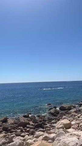 The video shows a rocky shoreline with the ocean in the background. The sky is clear and blue. There are three personal watercraft, each with at least one person on board, moving across the water.