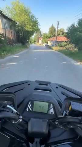 The media is a first-person perspective video recorded while riding a black ATV on a paved road through a village. The dashboard of the ATV is prominently visible, displaying a speed of 44 and then 45, indicating the vehicle is in motion. The road is lined with houses on both sides, some with red-tiled roofs. Trees and vegetation are abundant, suggesting a rural setting. In the background, there are a few parked cars. The sky is clear and blue, with bright sunlight casting shadows, indicating it is daytime, likely late morning or afternoon. The overall mood appears to be one of leisurely exploration or travel through a quiet, sunny village. The location is identified as Orlovtsi, Bulgaria.
