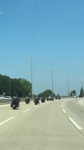 A group of motorcycles and cars are driving on a highway. The sky is blue and clear. There are trees and streetlights along the side of the road. The motorcycles are in the left lane, and the cars are in the right lane. The motorcycles appear to be riding in formation. The video is shot from a vehicle behind the motorcycles.