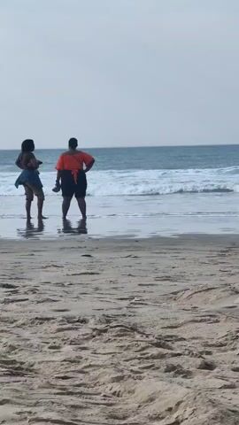 Two individuals are standing at the edge of the ocean on a sandy beach. The person on the left is wearing a blue top and what appears to be shorts or a skirt. They are facing away from the camera and towards the water. The person on the right is wearing an orange top and dark shorts, also facing away from the camera. Both individuals have their feet in the shallow water. The ocean is visible with waves rolling in towards the shore. The sky is overcast. The foreground is a sandy beach.